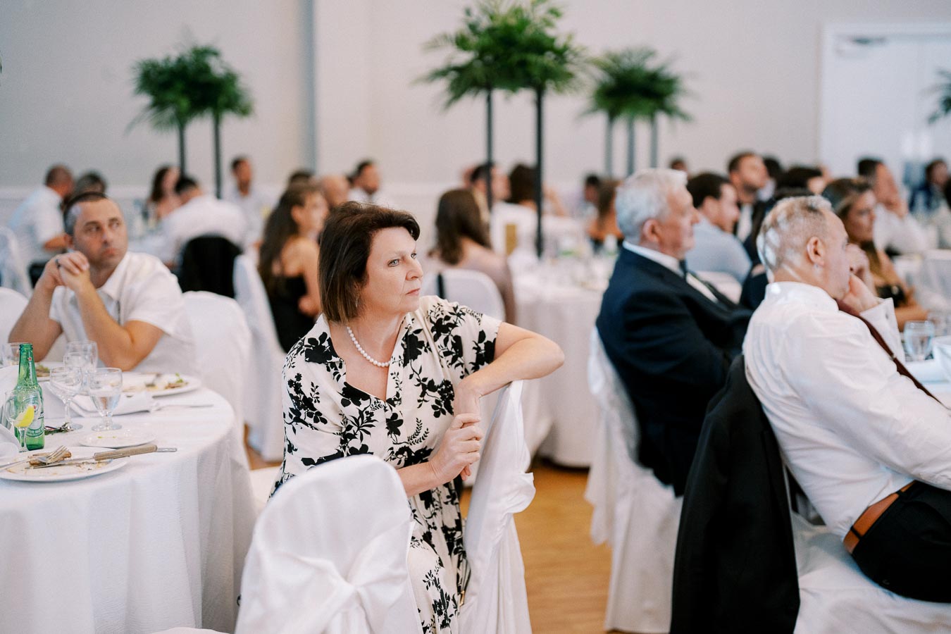 Guests attentively listening during a formal event, with elegantly decorated tables and lush green plants in the background, creating an atmosphere of sophistication and engagement.