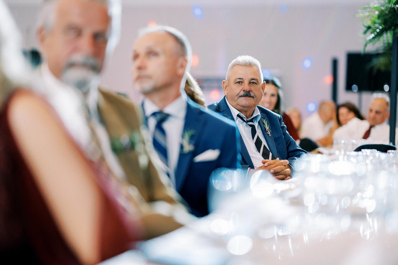 Senior man in a blue suit attentively listening at a formal event, surrounded by other guests.