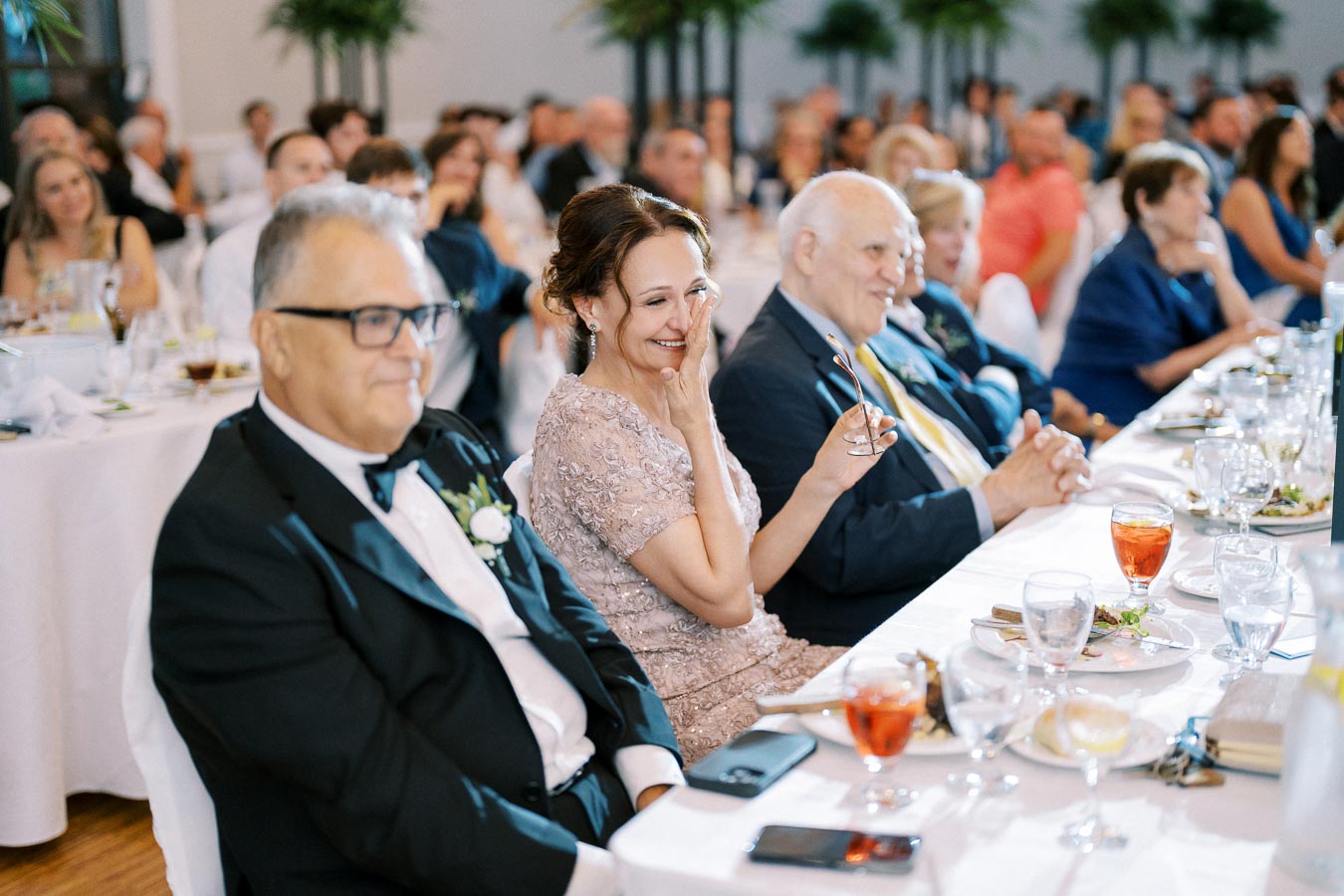 Guests enjoying a joyful moment at a formal event, seated at a decorated table, with glasses of beverages and plates of food, amidst a large gathering in a banquet hall.