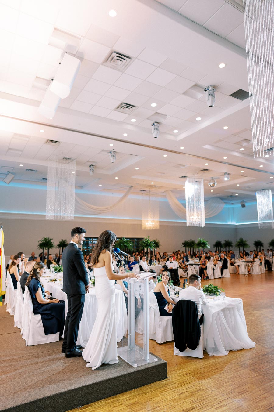 Wedding reception speech in elegant ballroom with guests seated at decorated tables, bride in white gown at podium, and ambient lighting with modern chandeliers.