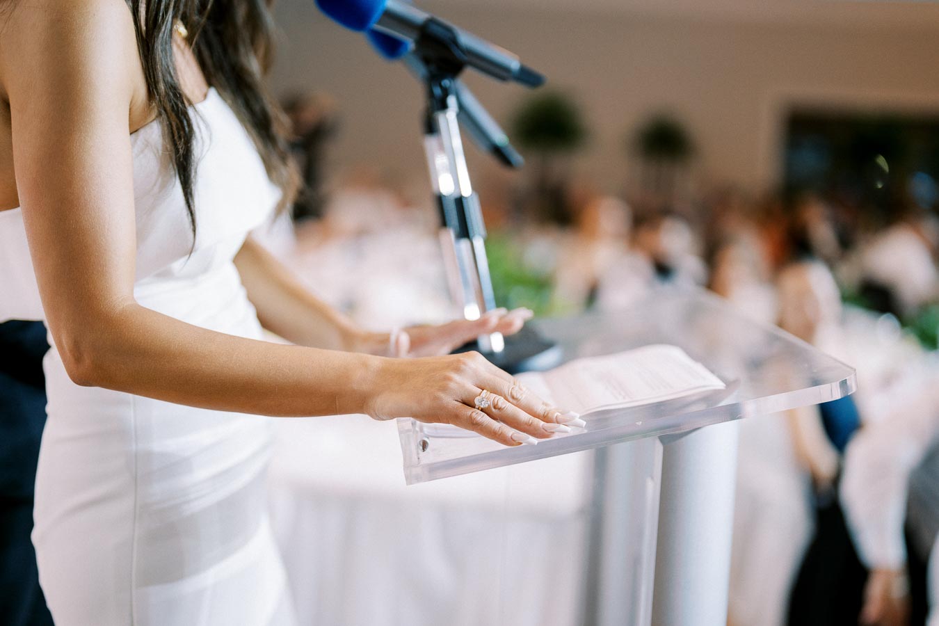 A speaker in a white dress gives a speech at a podium, holding a microphone, in front of a blurred audience during an event.