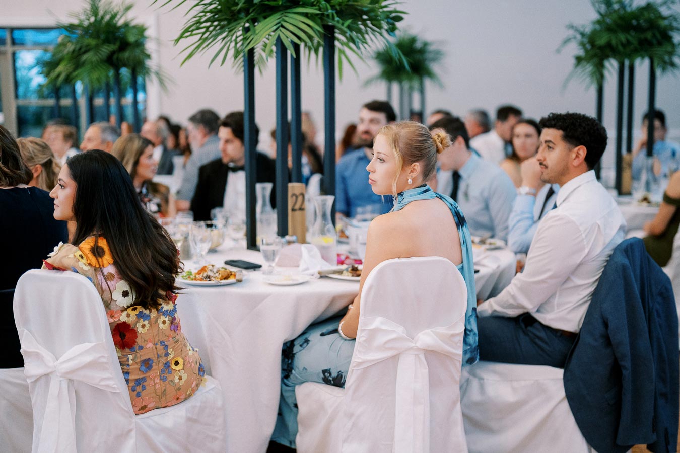 Guests seated at a formal event, wearing elegant attire and surrounded by table decorations, including tall green plants and white tablecloths.