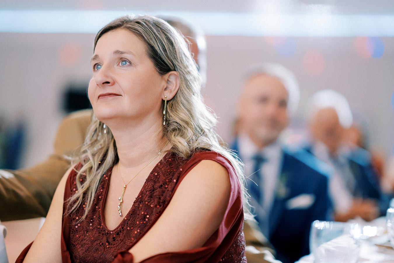 A woman in a red dress attentively watching an event indoors, with blurred people in the background, conveying a sense of focus and engagement.