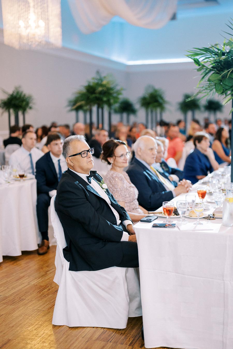 Guests sitting attentively at a formal event, with tables adorned with white tablecloths and glasses, surrounded by elegant decor and plants.