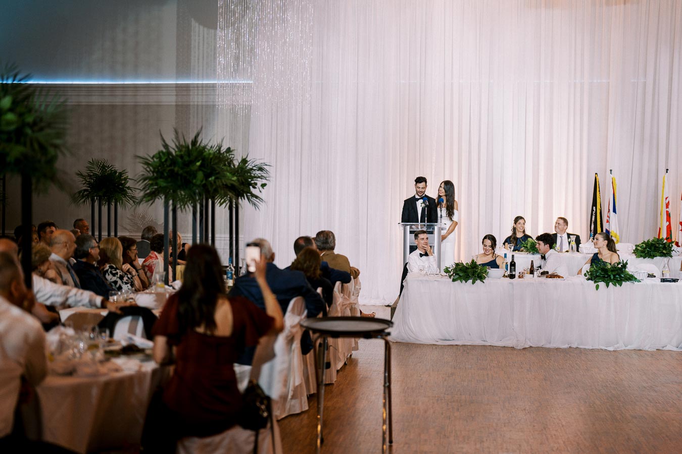 Elegant wedding reception with guests seated at round tables, featuring a couple delivering a speech at a podium adorned with white drapery in the background and greenery decorations on the tables.