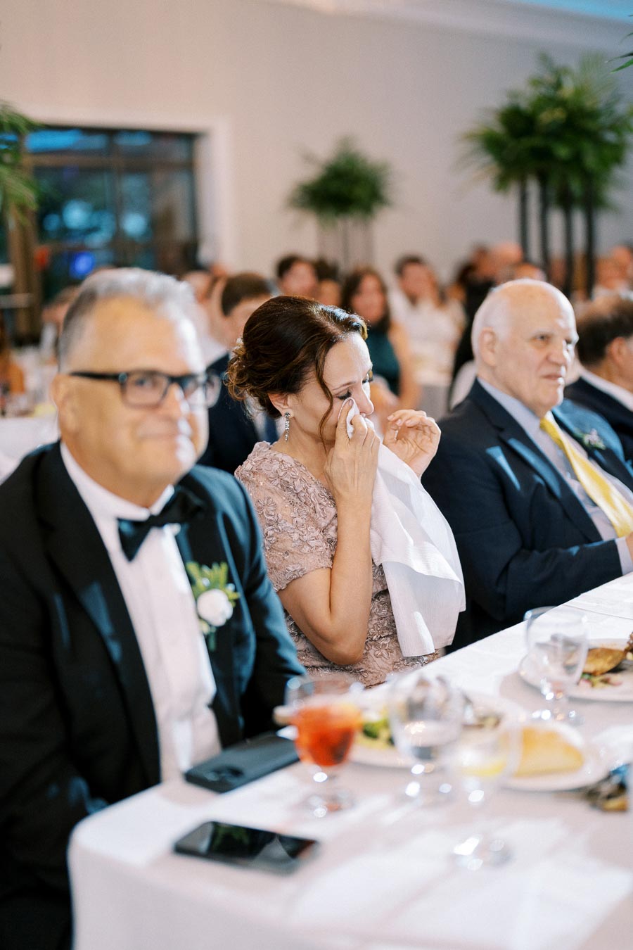 A wedding guest in formal attire wipes away tears of joy during a heartfelt moment at the reception, surrounded by other seated attendees in a beautifully decorated venue.