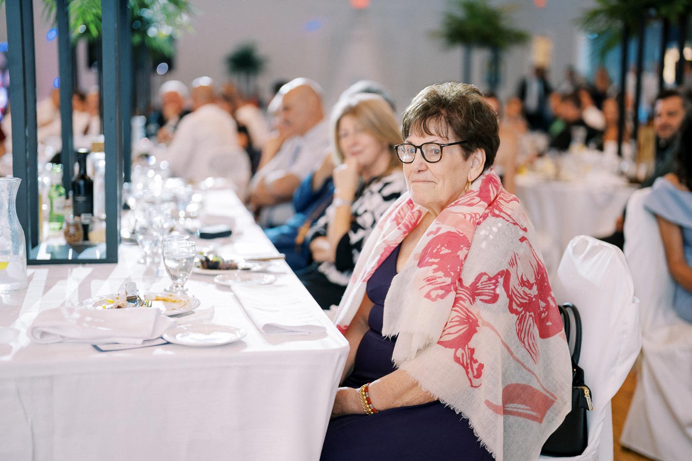 Elderly woman in glasses and a floral scarf sitting at a formal event, surrounded by a crowd of people in a banquet setting with white tablecloths and glassware.