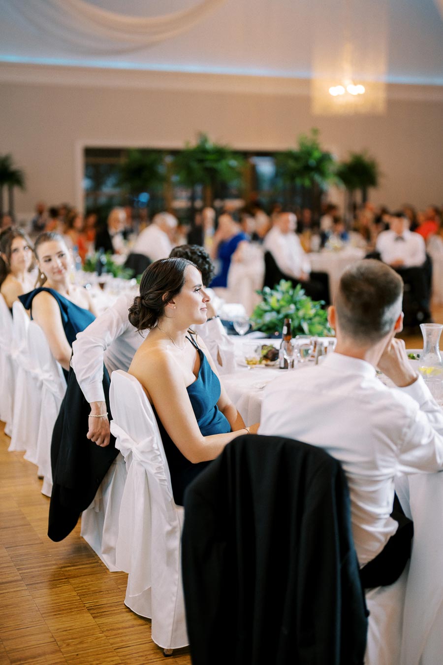 Elegant wedding reception with guests seated at tables adorned with white linens and lush green centerpieces, dressed in formal attire, attentively watching an unseen event.