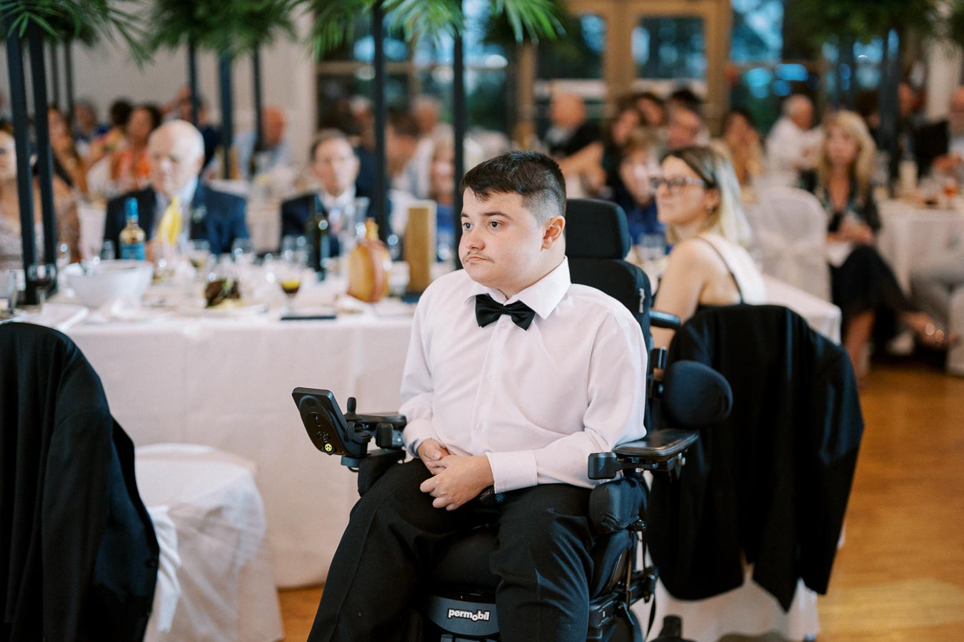 A young man in a wheelchair wearing a white shirt and bow tie at a formal event, surrounded by elegantly dressed guests seated at tables with white tablecloths and decorative plants.