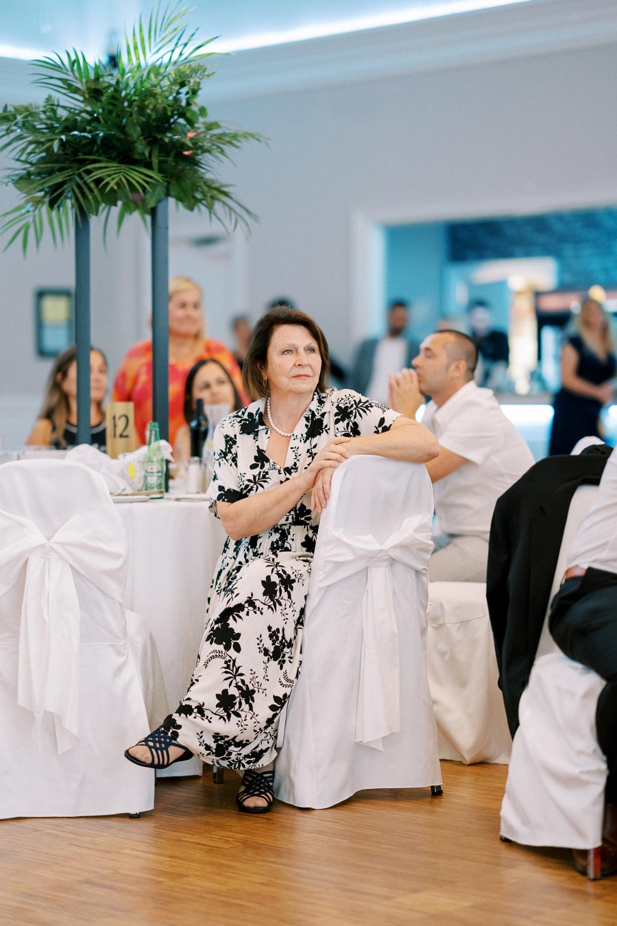 An elegantly dressed woman seated at a decorated table at a formal event, surrounded by other well-dressed attendees. The room features chic floral arrangements and ambient lighting, creating a sophisticated atmosphere.