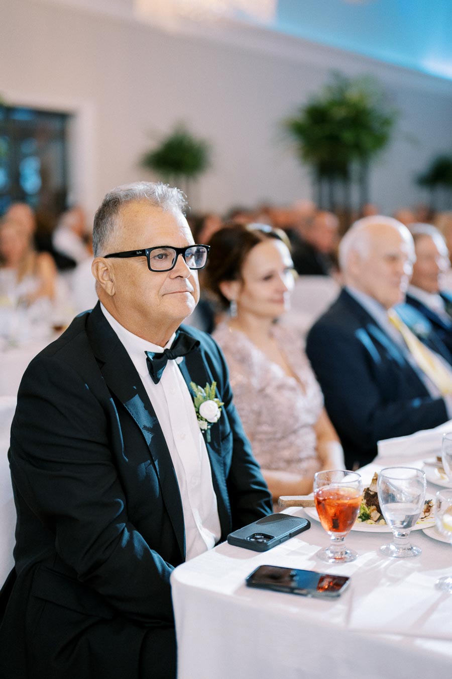 Elegant wedding guest wearing a tuxedo and glasses, sitting at a table with a festive drink and meal in a banquet hall setting.