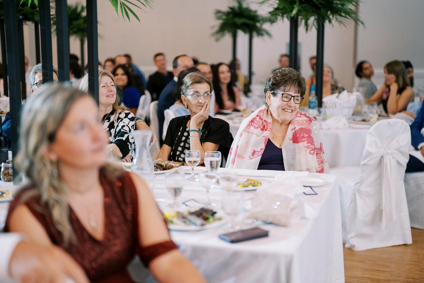 A diverse group of people sitting at round banquet tables with white linens, attentively watching a speaker, surrounded by elegant greenery in a formal event setting.