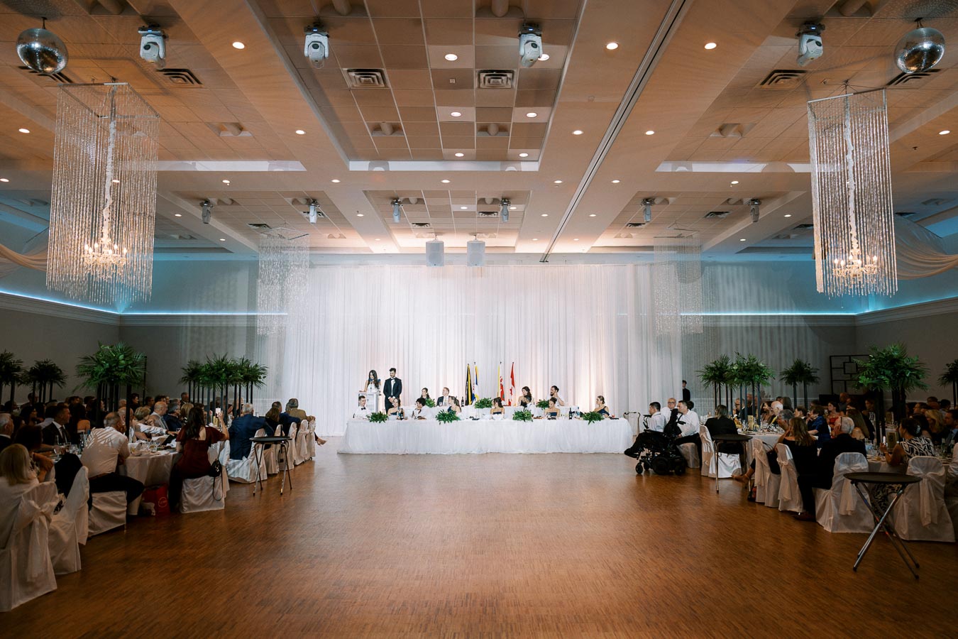 Elegant wedding reception in a spacious ballroom with guests seated at tables, a long head table with a couple standing at the podium, adorned with white drapery and greenery, under modern chandeliers.