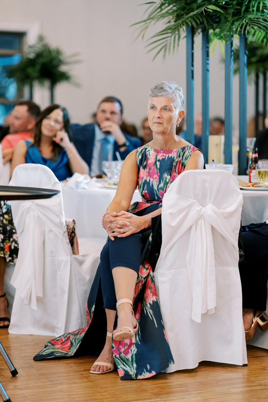 Elegant elderly woman in floral dress seated at a formal event, surrounded by guests in a decorated banquet hall.