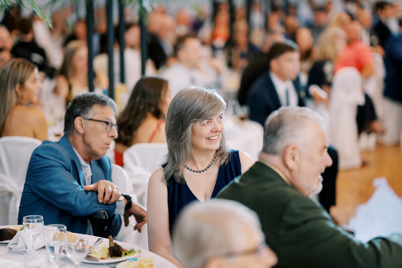 Smiling woman with gray hair in a blue dress seated at a formal event, surrounded by other attendees at tables, enjoying food and conversations.