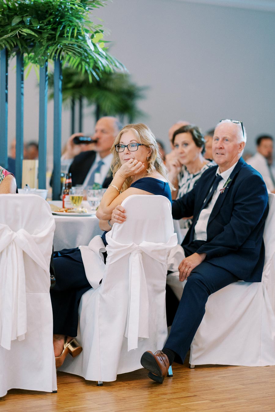 Guests attentively seated at a formal event with elegantly decorated chairs and lush indoor plants.