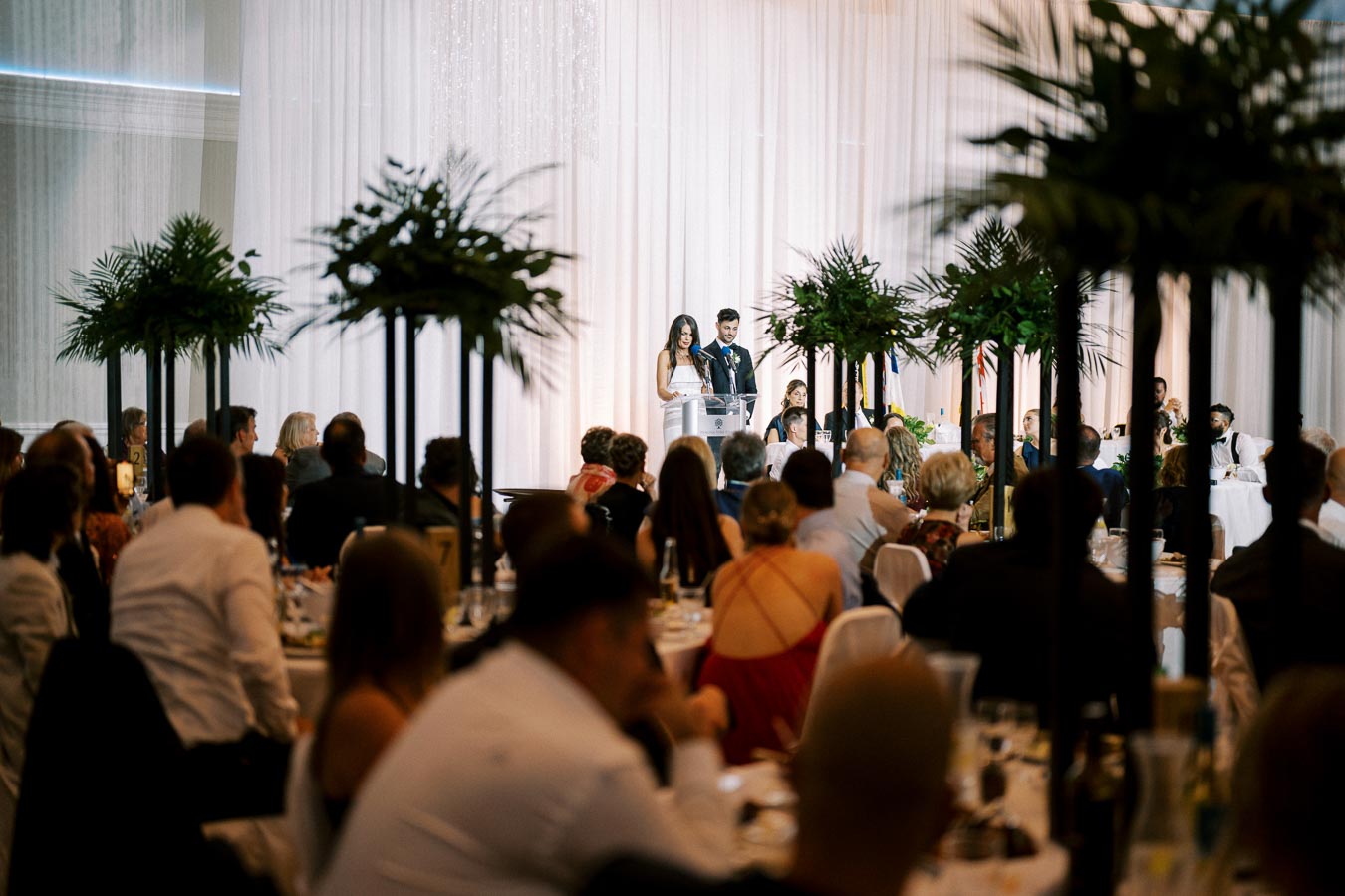 A well-dressed couple delivers a speech at a formal event, surrounded by an elegantly decorated venue with lush greenery and seated guests attentively listening.