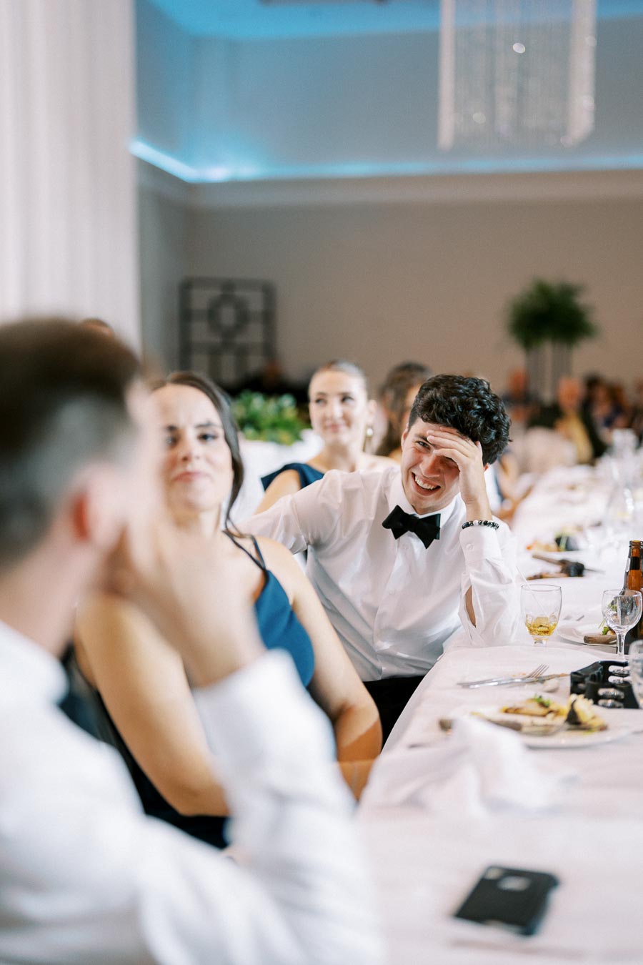 Group of people enjoying a formal dinner event; man in white shirt and black bow tie smiling, seated at a decorated table with drinks and tableware.