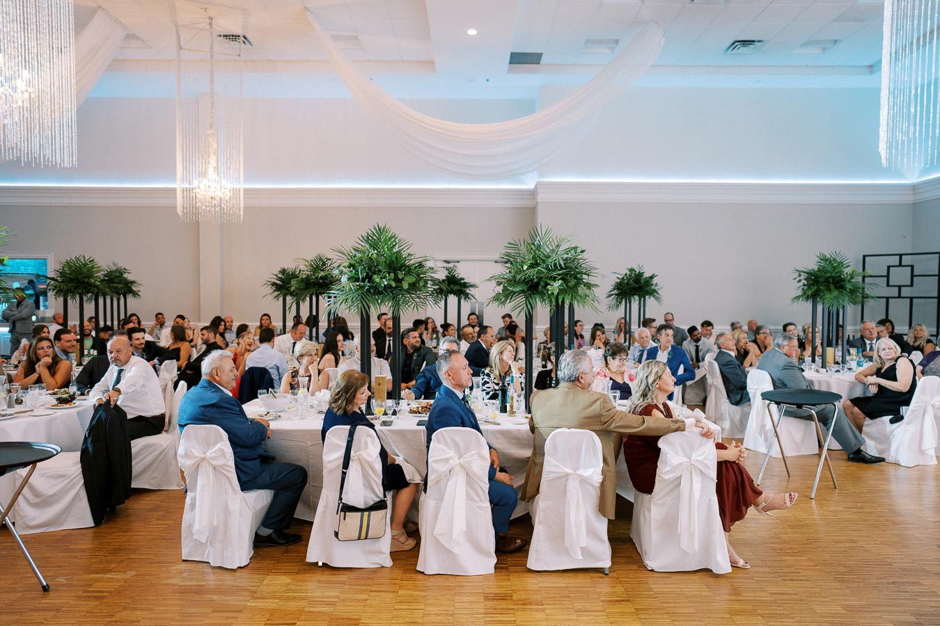 A large group of elegantly dressed guests seated at round tables, attentively watching a presentation in a beautifully decorated banquet hall with tall greenery centerpieces and white draped ceilings, creating a sophisticated event atmosphere.