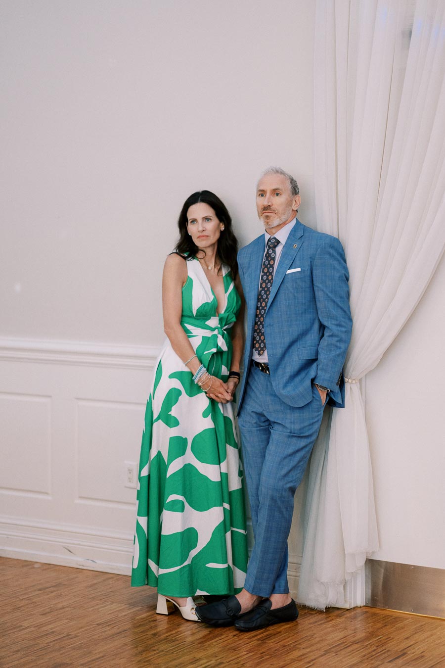 Man in a blue suit and woman in a green and white patterned dress standing together, leaning against a white wall with curtains, wooden floor background.