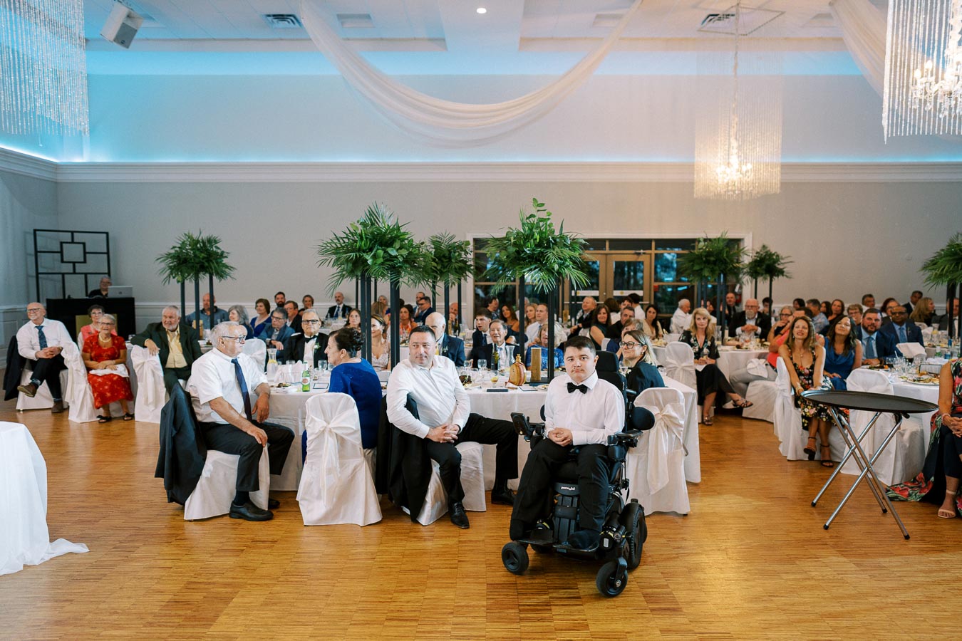 A large group of people dressed in formal attire seated at decorated tables in an elegant banquet hall, featuring white linens and tall green centerpieces, attentively watching an event unfold.