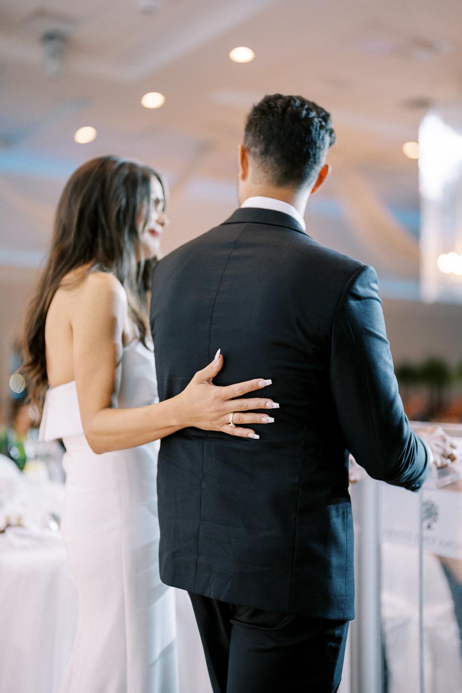 Elegant wedding couple sharing a moment, with the bride in a stylish white gown gently embracing the groom in a black suit at a beautifully decorated indoor venue.