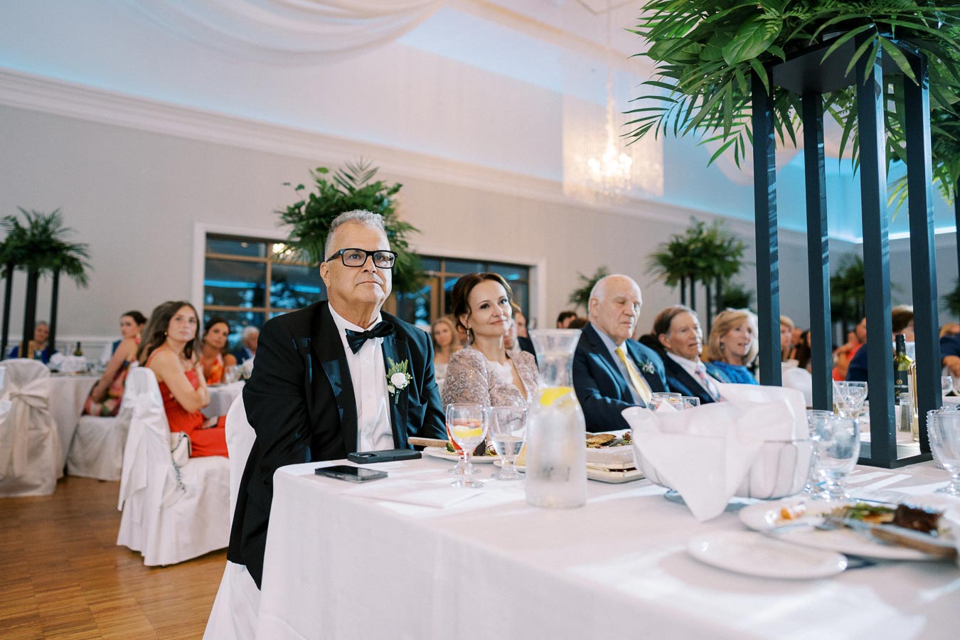 People seated at a wedding reception, attentively listening to a speech. The elegantly decorated room features potted plants, white tablecloths, and a chandelier.