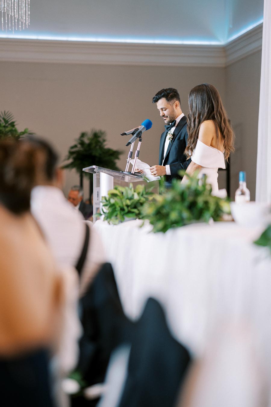 A couple delivering a speech at a wedding reception, standing at a podium with a blue microphone. The room is elegantly decorated with green plants and white floral arrangements.