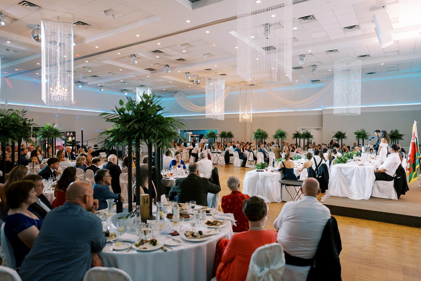 Wedding reception in a beautifully decorated banquet hall, featuring elegantly dressed guests seated at round tables, lush greenery, and crystal chandeliers enhancing the sophisticated atmosphere. A speaker addresses the attendees from a well-decorated head table.