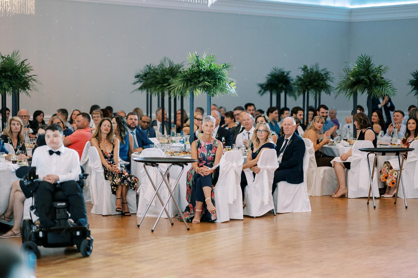 A diverse group of elegantly dressed people seated at a formal event, surrounded by decorative greenery and white table settings, attentively listening to a speech or presentation.