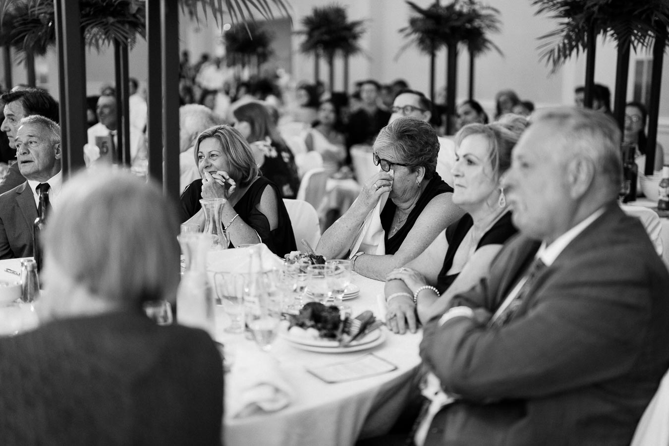 Black and white photo of a group of people attentively listening at a formal event, seated around a banquet table, with elegant decor and tableware.