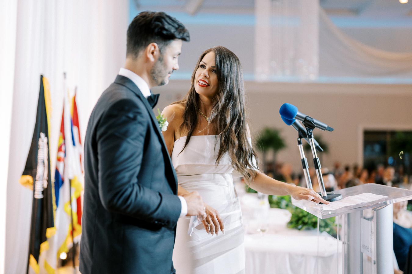 A man and woman in formal attire at a podium with a blue microphone, flags in the background, during a formal event or ceremony.