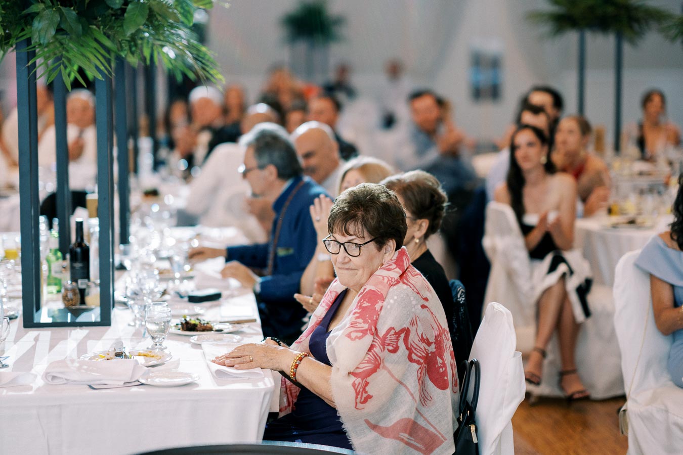 A group of people seated at elegantly decorated tables during a formal event, with a focus on an elderly woman in a pink and white shawl, surrounded by plates and drinks, suggesting a lively and social atmosphere.