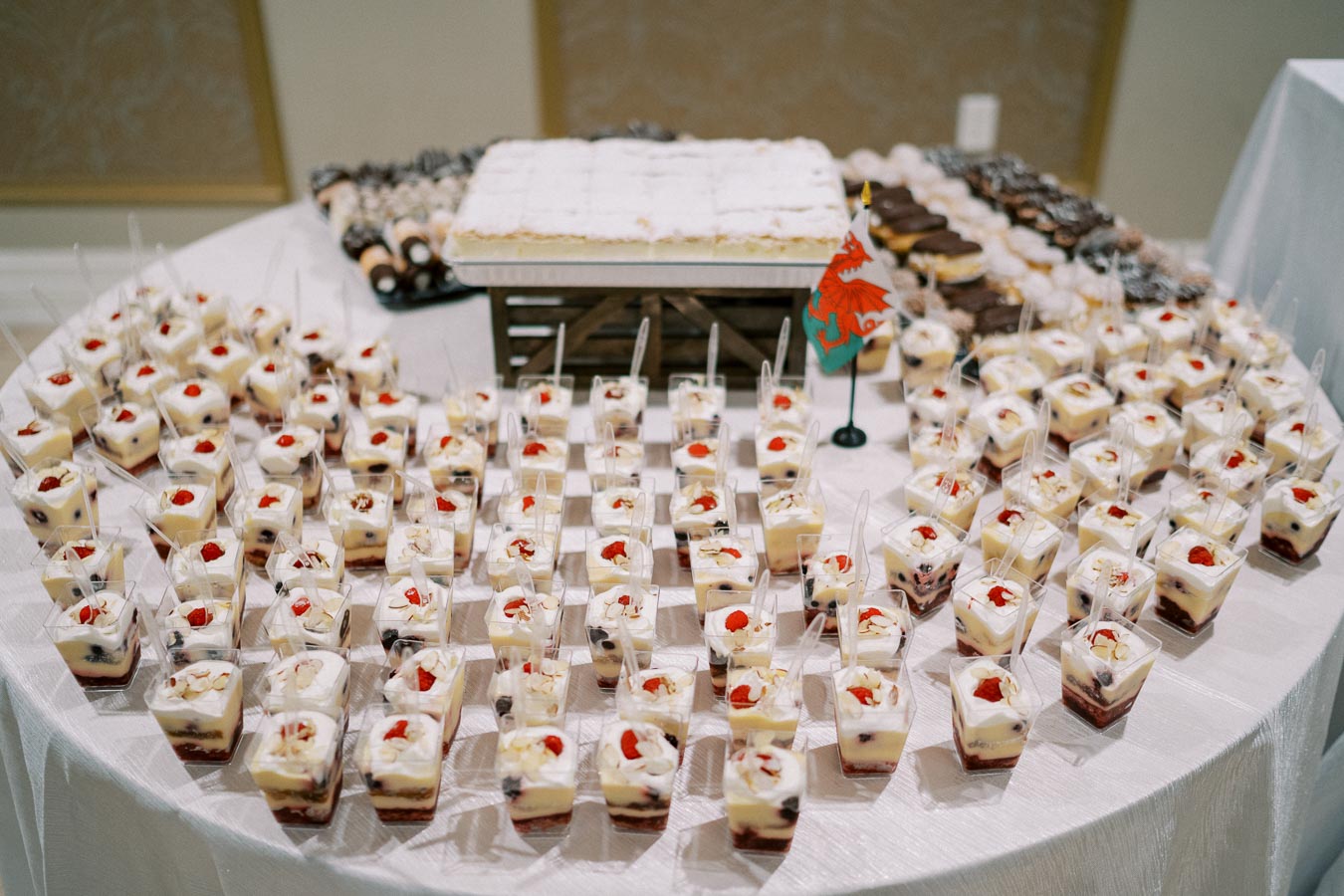 A decorative dessert table displaying an assortment of layered parfaits in small cups, topped with cherries and whipped cream, alongside other pastries, ideal for a buffet or catered event.