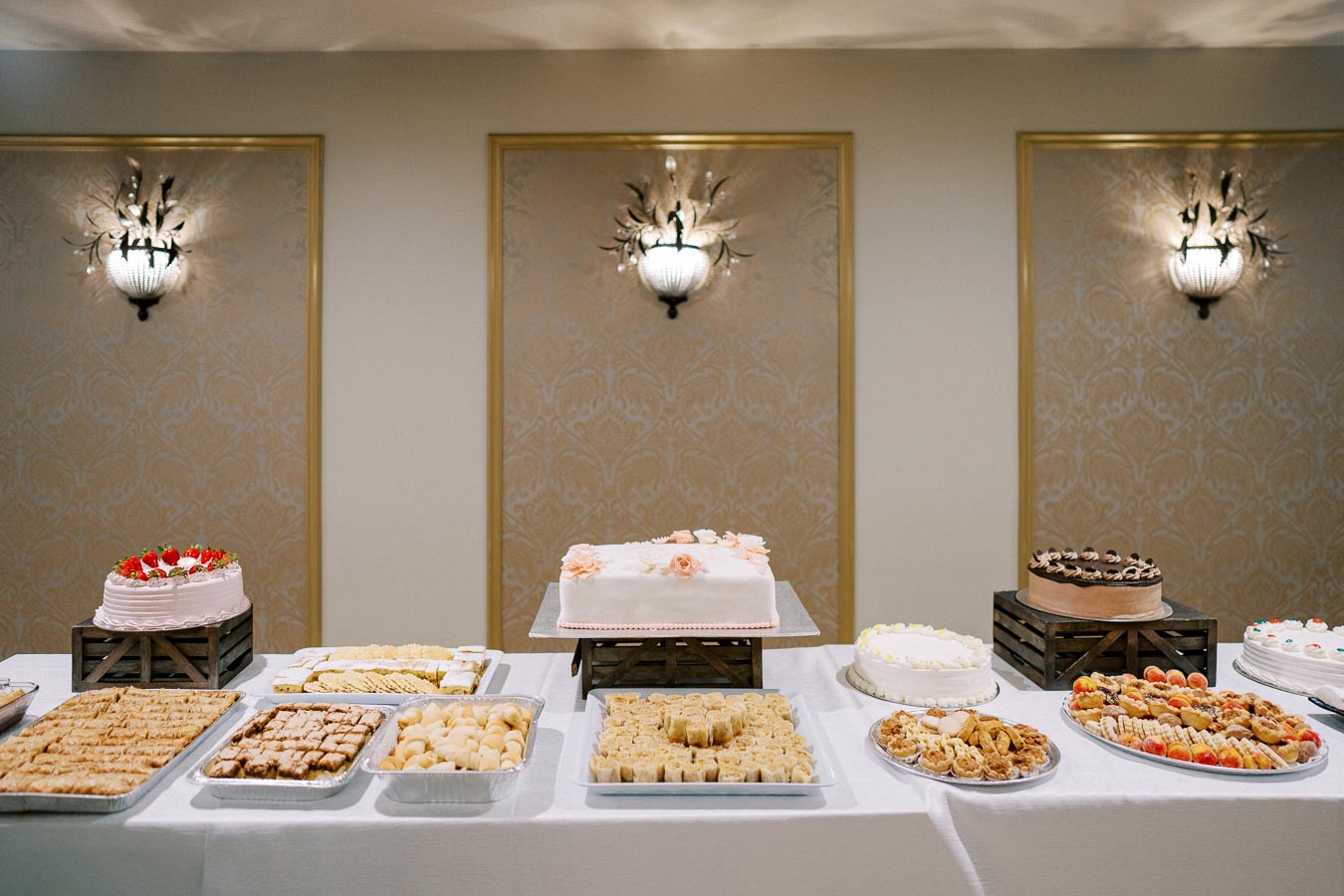 Dessert buffet table with various cakes and pastries in a well-lit banquet hall, featuring decorative wall sconces and ornate wallpaper.