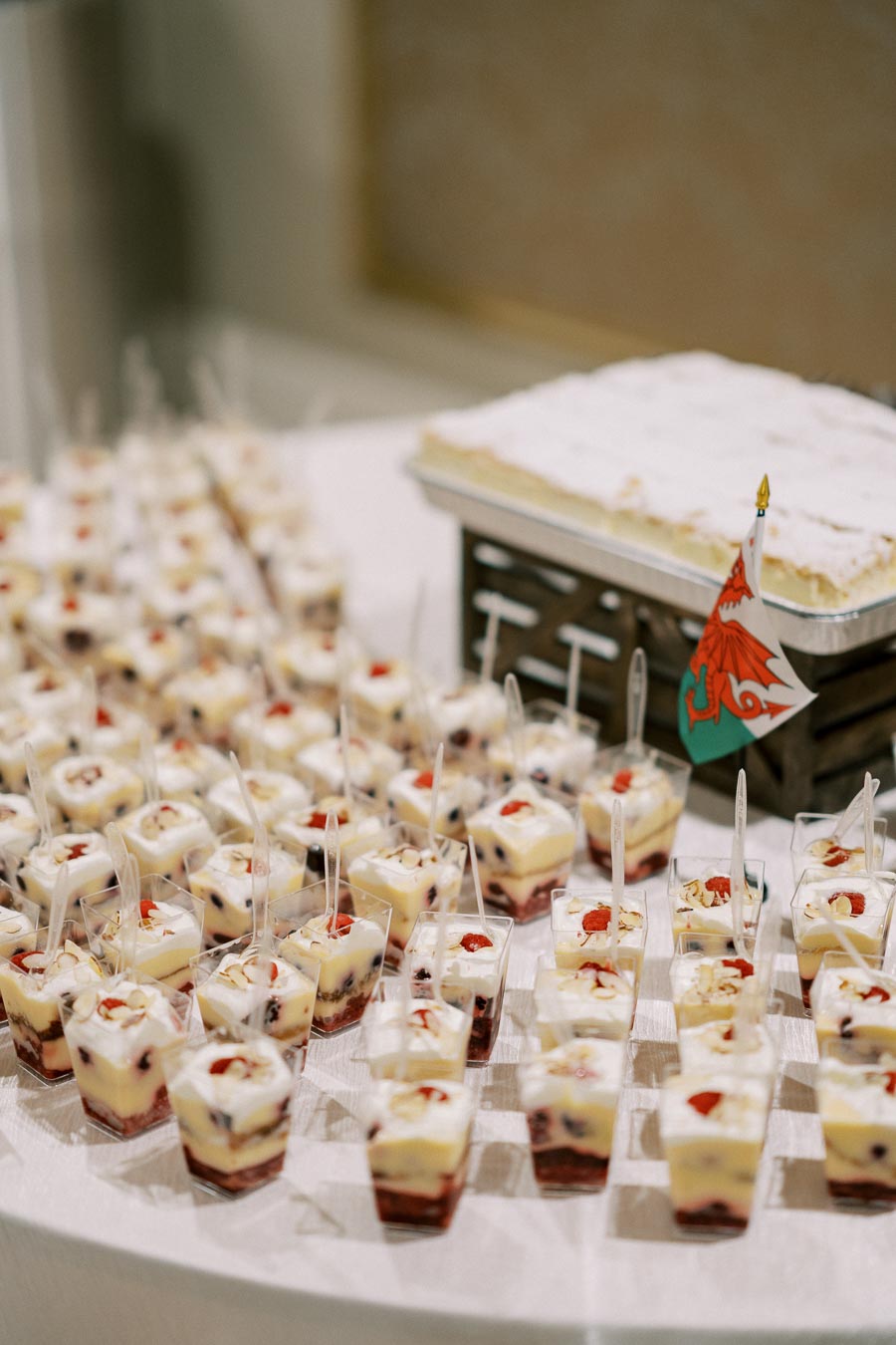 Individual servings of layered dessert cups on a table, featuring a creamy topping with fruit garnishes, next to a large dessert tray decorated with a festive flag.