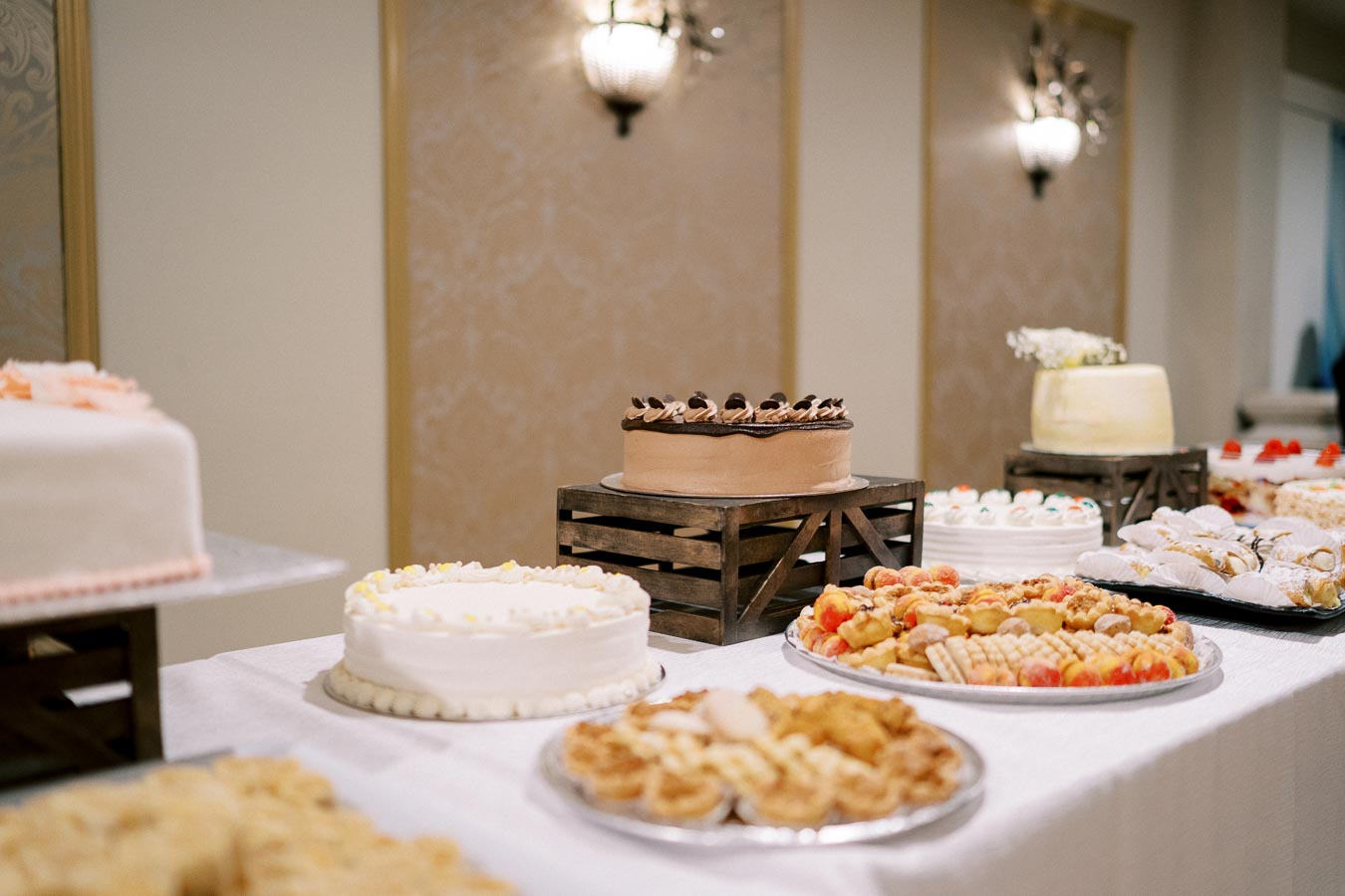 An elegant dessert table at an event featuring a variety of cakes and pastries, including a chocolate cake centerpiece, surrounded by assorted treats on a white tablecloth in a warmly lit room.