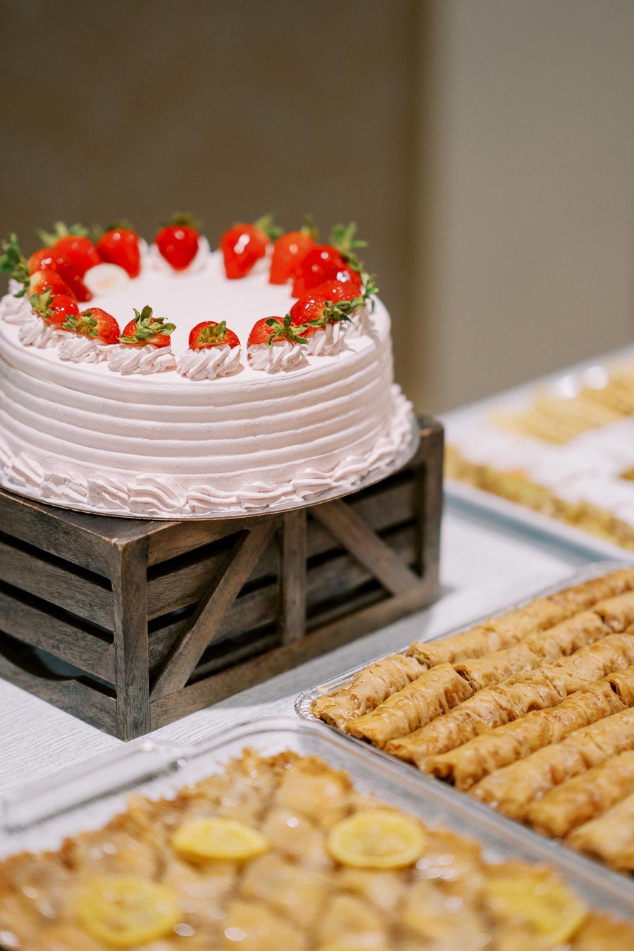 Strawberry-topped cake on a wooden stand alongside trays of baklava and pastries on a dessert table.