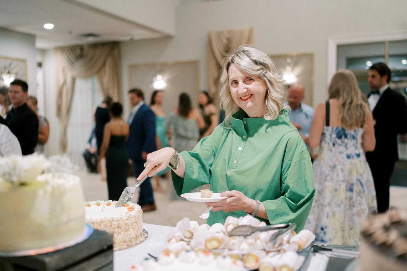 A woman in a green blouse smiling and serving cake at a social event, with a blurred background of people mingling in formal attire.