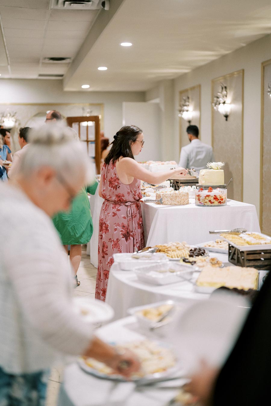 Guests serving themselves at a buffet table during a wedding reception, with a variety of desserts and savory dishes displayed. The setting includes elegantly decorated walls and ambient lighting, suggesting a formal event.