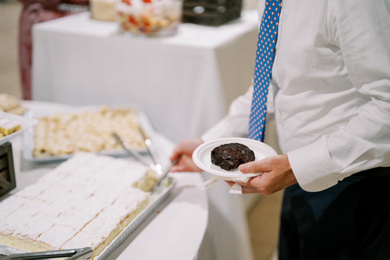 A person in a white shirt and patterned tie selects a dessert at a buffet, holding a plate with a chocolate cake, with trays of pastries and other treats displayed on white tables in the background.