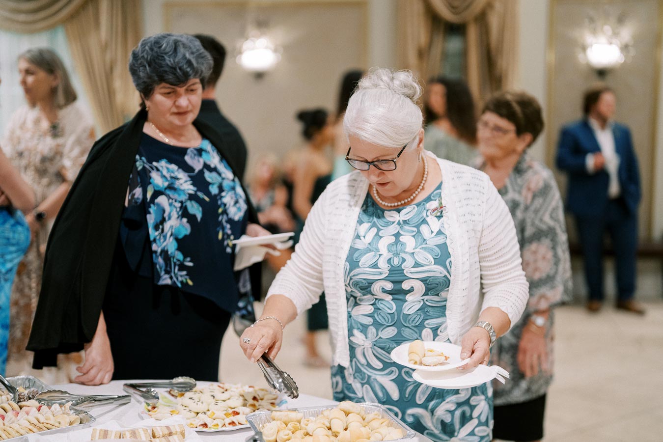 Elderly women selecting pastries from a buffet table at a social gathering, surrounded by other guests in formal attire.
