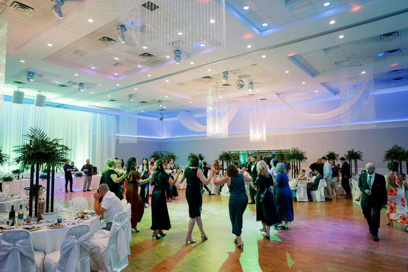 People dancing in a beautifully decorated banquet hall with elegant lighting and tables adorned with white linens and greenery.