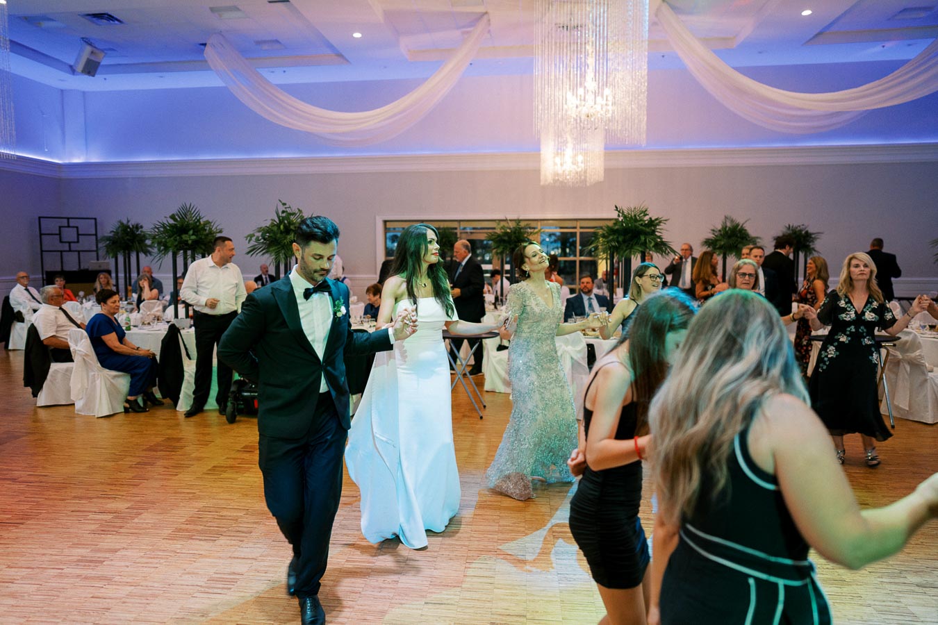 A lively wedding reception with guests dancing joyfully on a polished wooden floor. The bride in a white gown dances with a groom in a black tuxedo, surrounded by elegantly dressed attendees. The venue is beautifully decorated with draped fabric and a crystal chandelier, adding a touch of elegance to the celebration.