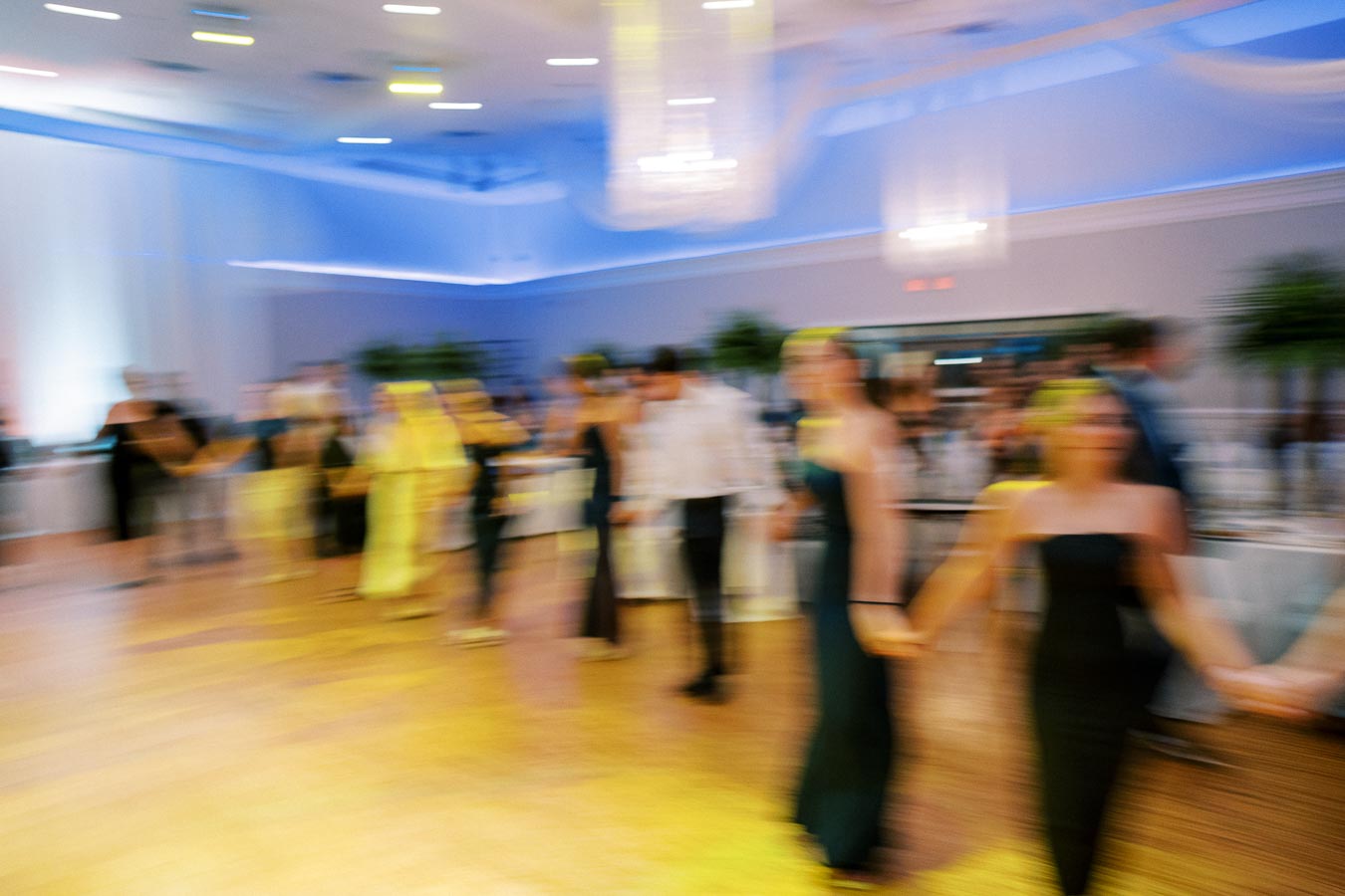 Blurred image of people holding hands while dancing in a well-lit reception hall, conveying movement and celebration.