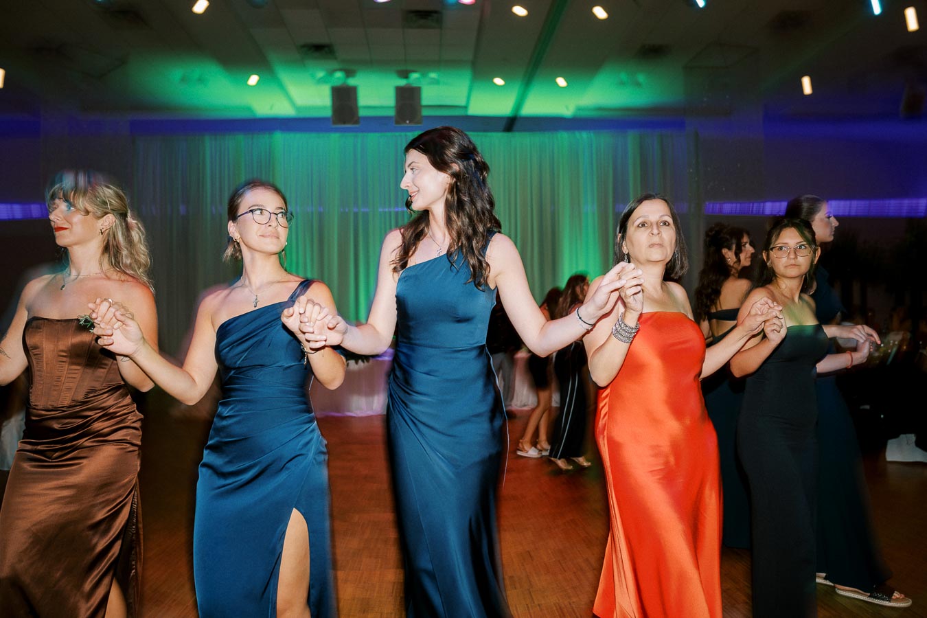 A group of women in elegant dresses dancing together at a formal event, with colorful lighting and a lively ambiance.