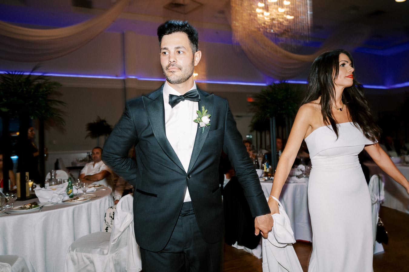 A elegantly dressed couple in formal attire entering a beautifully decorated wedding reception hall, with the man wearing a black tuxedo and the woman in a white evening gown, surrounded by elegantly set tables and ambient lighting.