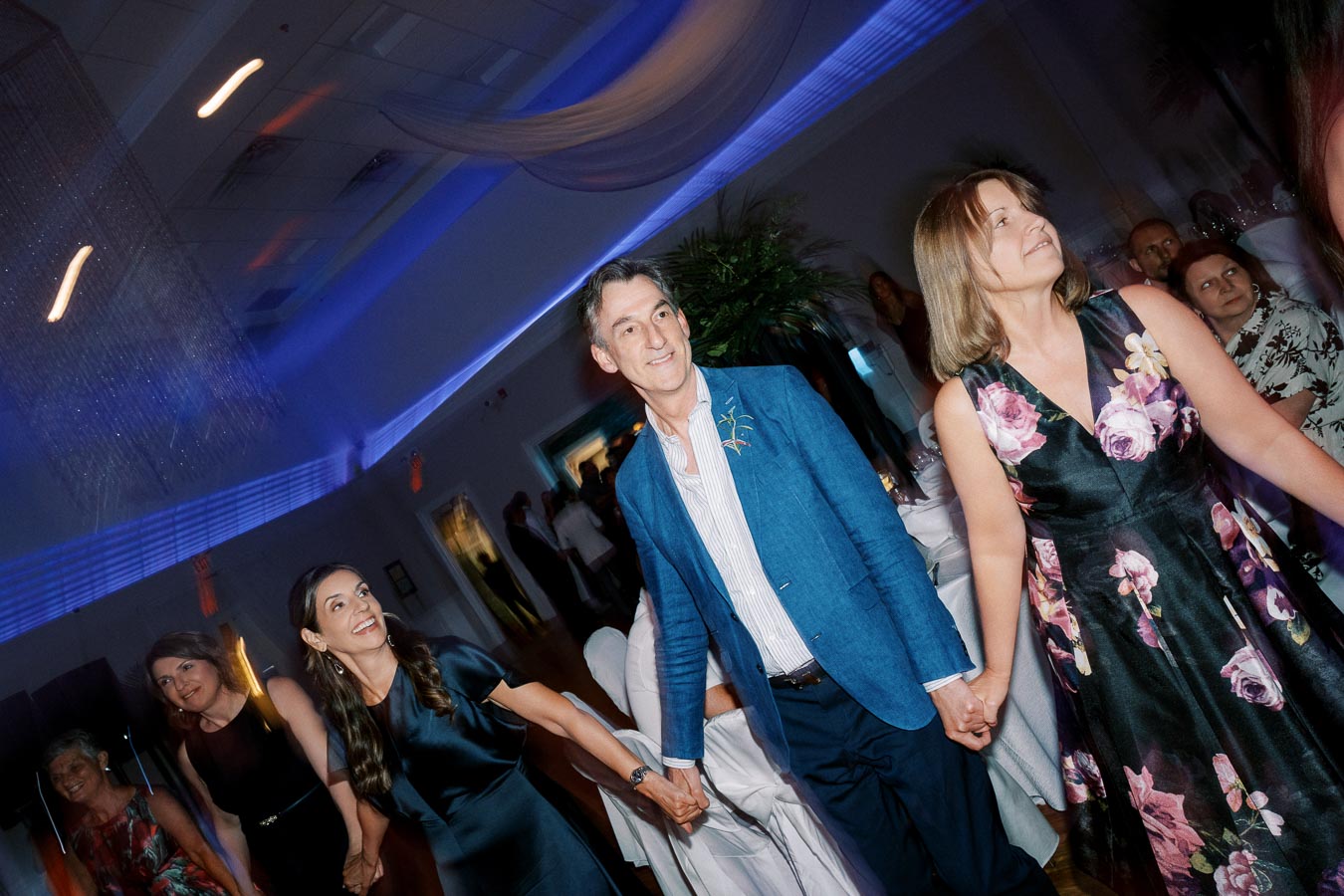 A group of people joyfully dancing and holding hands at a lively celebration, surrounded by elegant blue lighting and festive decor.