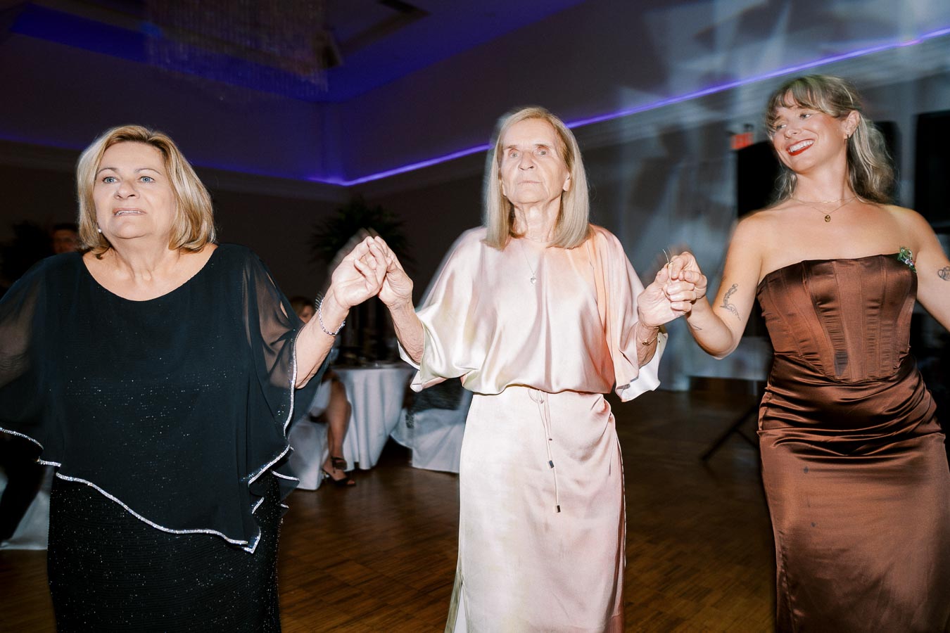 Three women joyfully holding hands and dancing at an indoor event, dressed in elegant evening attire.