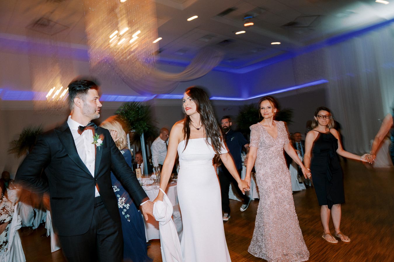 Elegant wedding celebration with a bride in a white gown and guests in formal attire dancing in a beautifully lit reception hall.